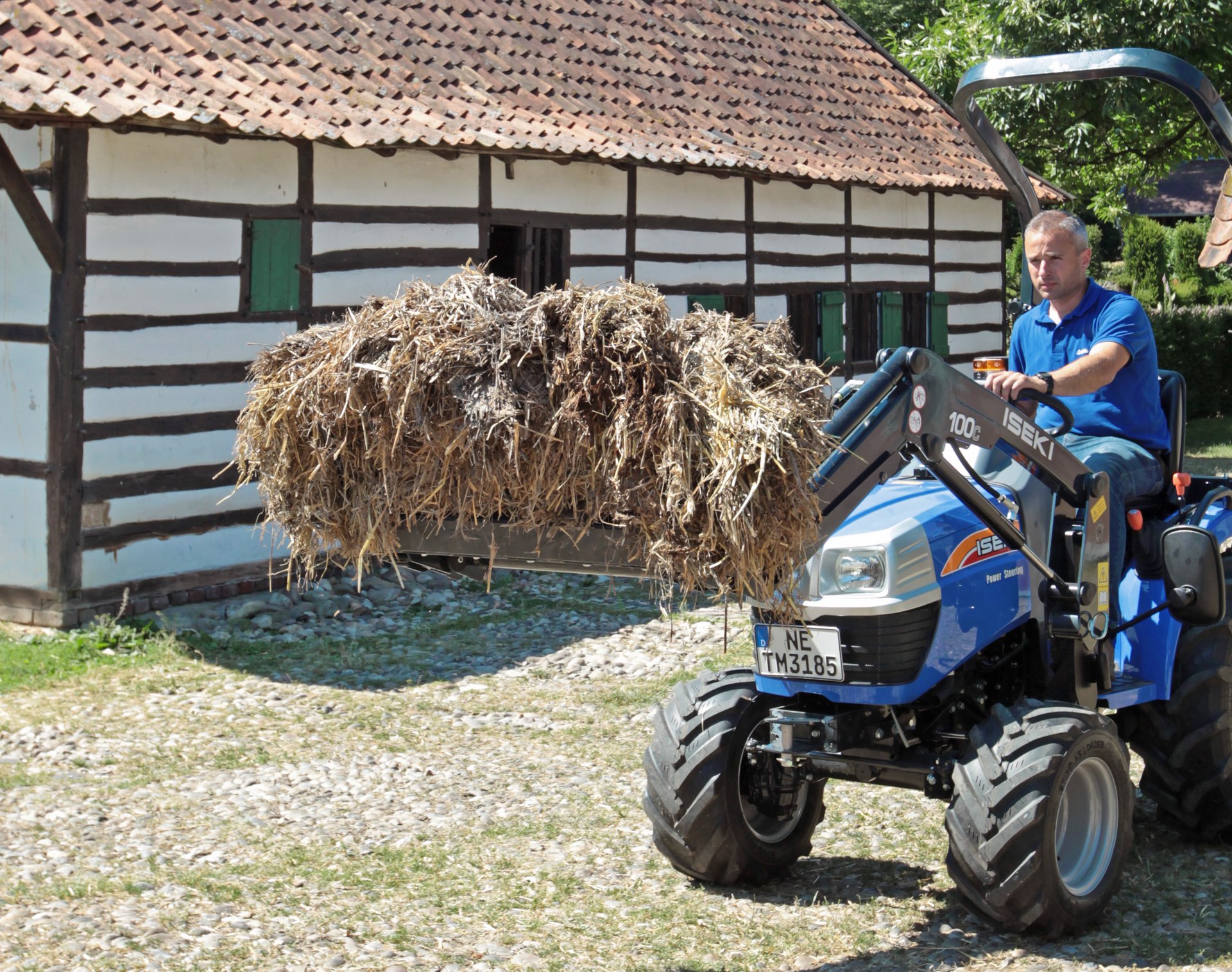 Ein ISEKI-Traktor mit Heu in der Schaufel wird über die Wiese vor einem Bauernhaus gefahren. 